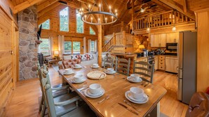 L’intérieur d’un chalet en bois comprend un coin repas, une cuisine, un mur en pierre et de grandes fenêtres.