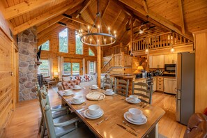 A wooden cabin interior with a dining area, kitchen, stone wall, and large windows.