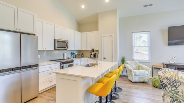 A modern kitchen with white cabinets, a stainless steel refrigerator, a microwave, and a large island with yellow bar stools. - Carolina Beach Vacation Home Near Boardwalk (Carolina Beach)