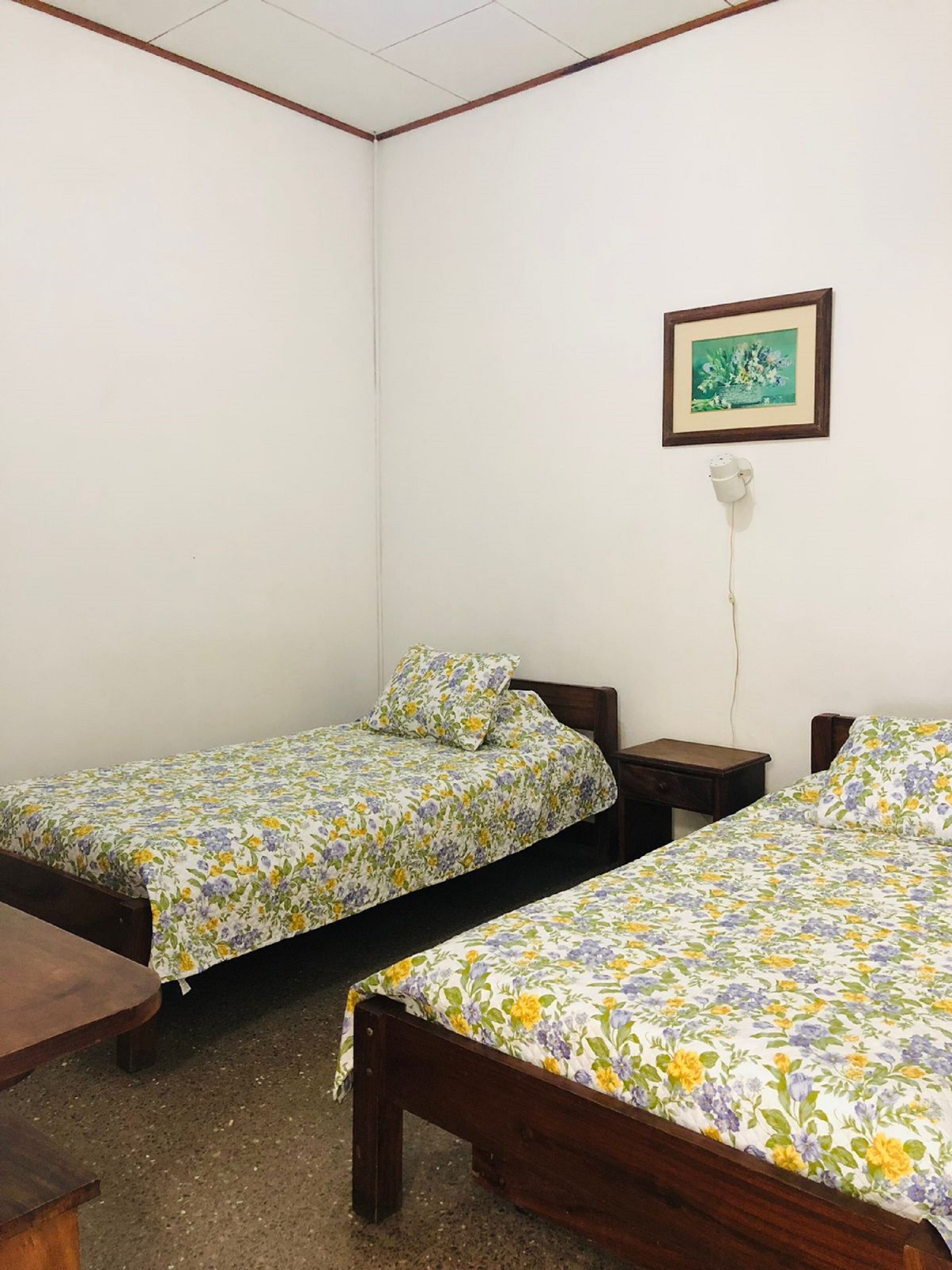 A room with two wooden beds, each with floral bedding, a small wooden nightstand, a framed picture on the wall, and a white electrical outlet.