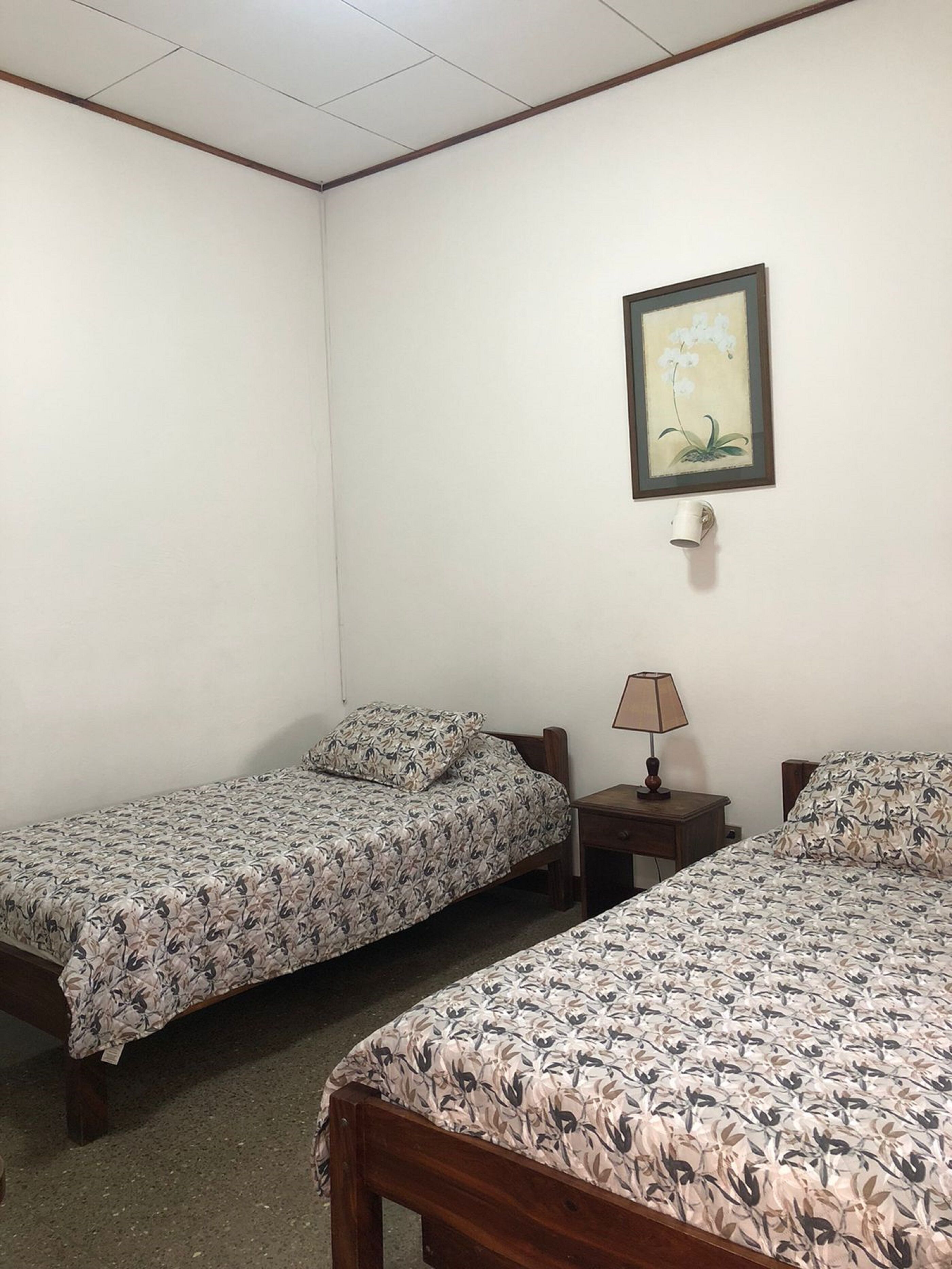 A room with two single beds, each with a wooden frame and floral-patterned bedding. There is a small bedside table with a lamp on one side and a framed picture on the wall above the bed on the other side.