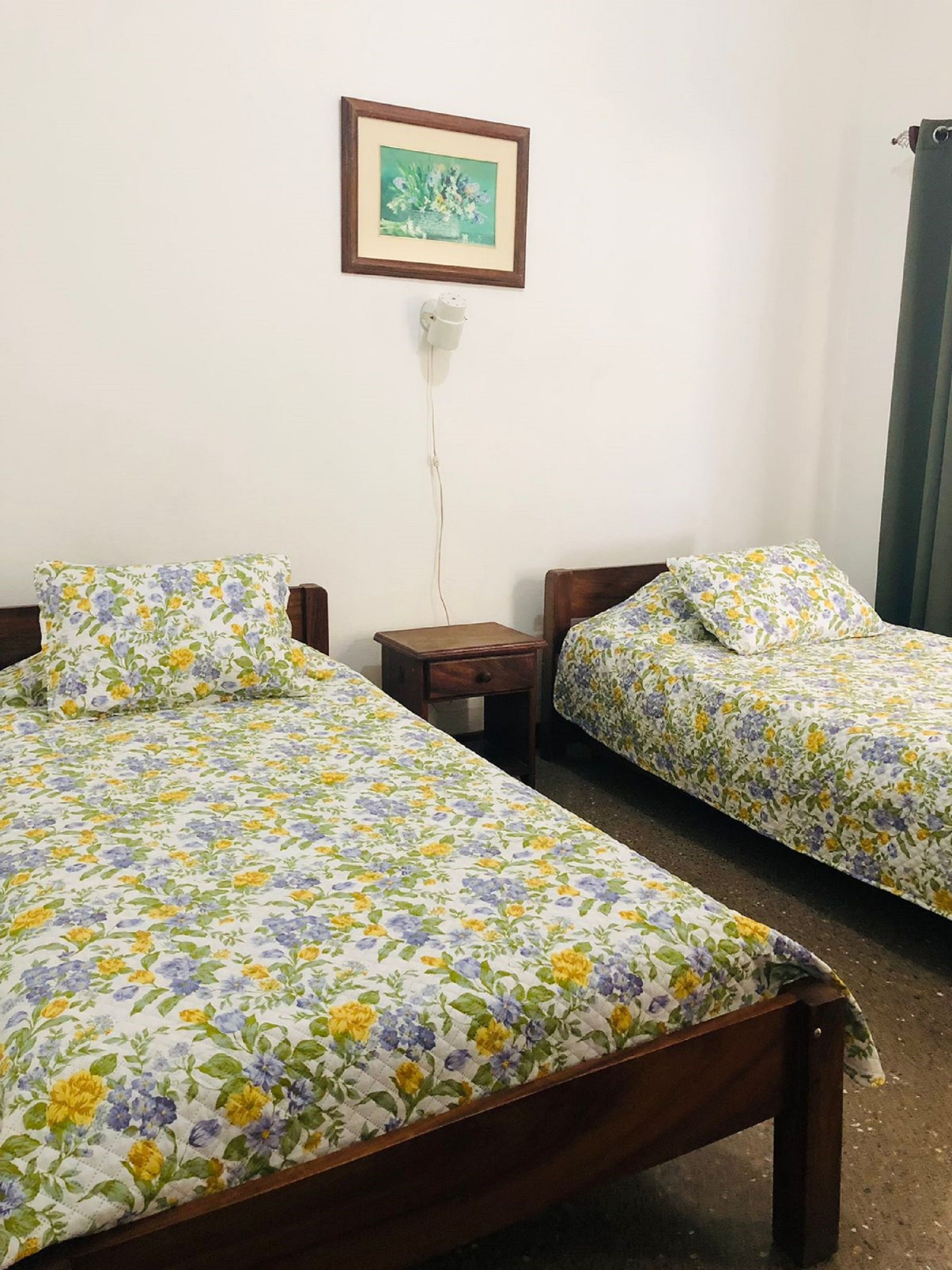 Een kamer met twee eenpersoonsbedden, elk voorzien van bloemenbeddelsels, een klein houten nachtkastje, een stopcontact op de muur en een lijst met een afbeelding erboven.