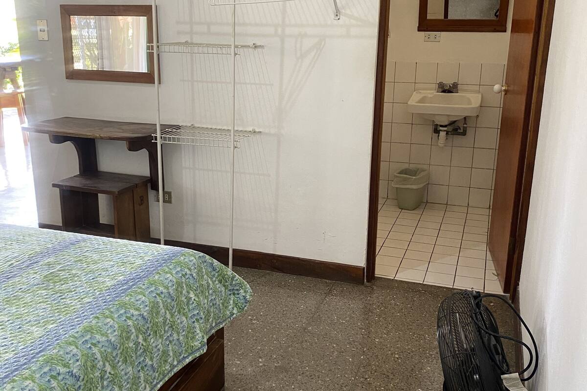 Une chambre comprenant un lit, un bureau, un étendoir fixé au mur, un miroir, un lavabo et un ventilateur.