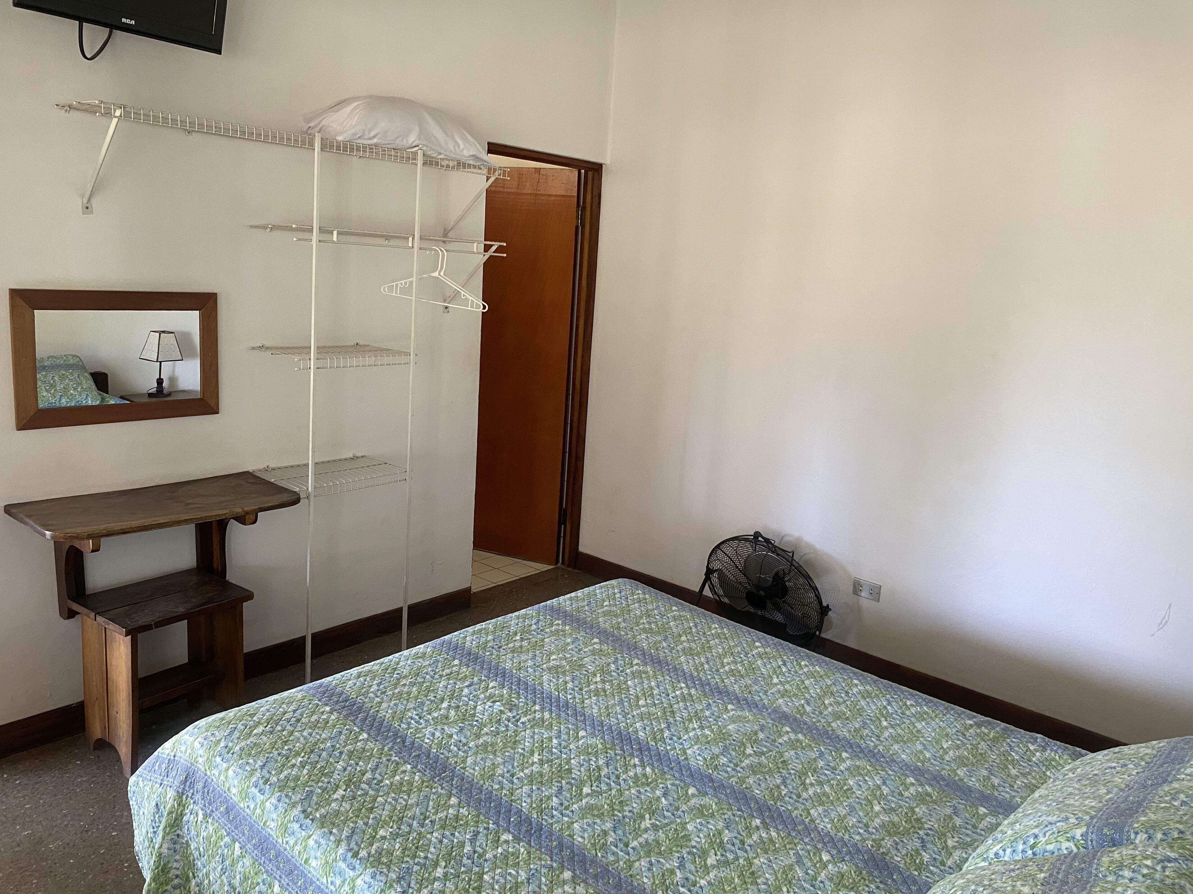 Une chambre à coucher comprenant un lit, une table de chevet, un miroir, une armoire et un ventilateur sur pied.
