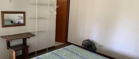 Une chambre à coucher comprenant un lit, une table de chevet, un miroir, une armoire et un ventilateur sur pied.