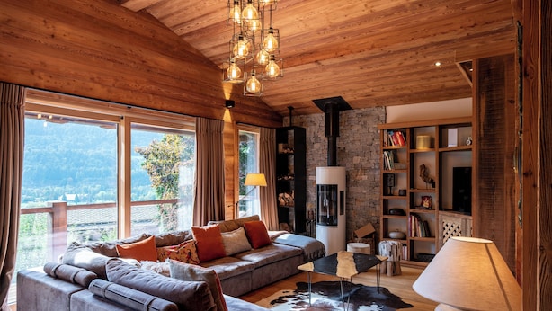 Ein gemütliches Wohnzimmer mit Kamin aus Stein, Holzboden und großen Fenstern mit Blick auf eine reizvolle Landschaft.