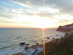 Ein Küsten-Sonnenuntergang mit einem Pier, der sich ins Meer erstreckt, Gebäuden auf einer Klippe und Felsformationen.