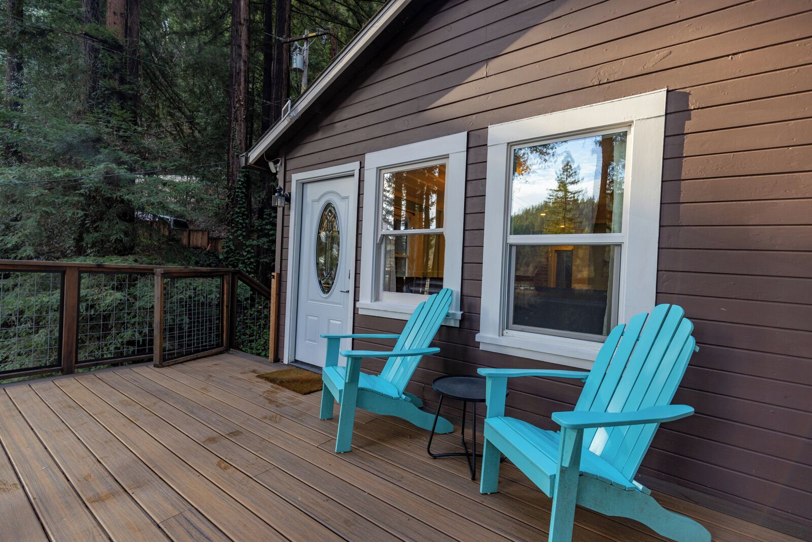 Een houten terras met twee turquoise Adirondack-stoelen en een klein tafeltje, met uitzicht op een bebost gebied.