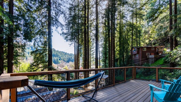 A wooden deck with a hammock and chairs, overlooking a forested area and a small building.