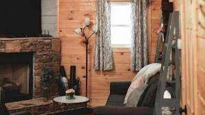 Interior acollidor d'una cabana amb una llar de pedra, parets de fusta, un ventilador de sostre i una finestra amb cortines.