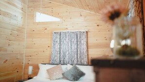 Habitació de cabana de fusta amb un llit, una tauleta de nit, una finestra i una cortina.