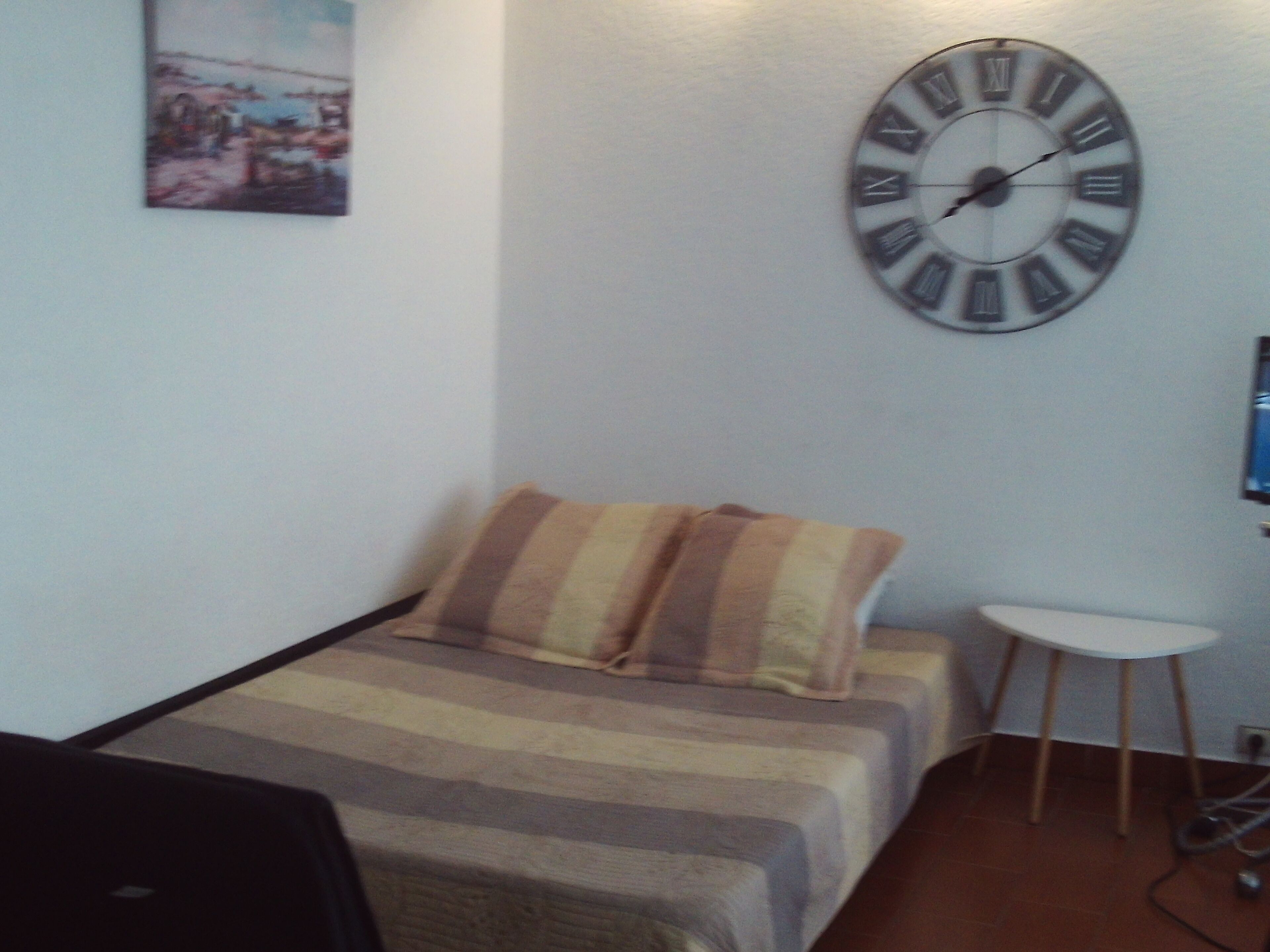 Een kleine kamer met een bed, een grote wandklok, een klein tafeltje en een schilderij aan de muur.