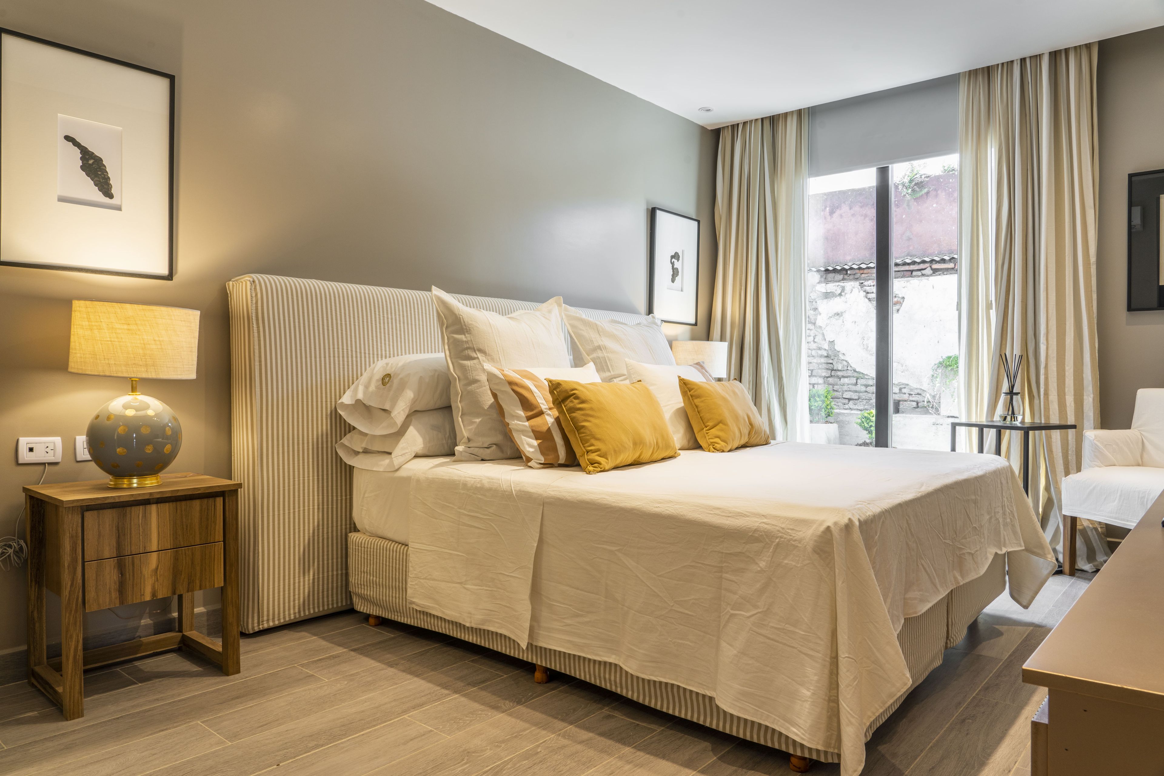 Habitación de hotel con una cama grande, una mesita de noche con lámpara, una silla y vistas al exterior.