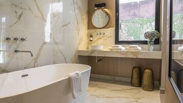 A modern bathroom with a freestanding bathtub, a round mirror, and a window with a view of greenery.