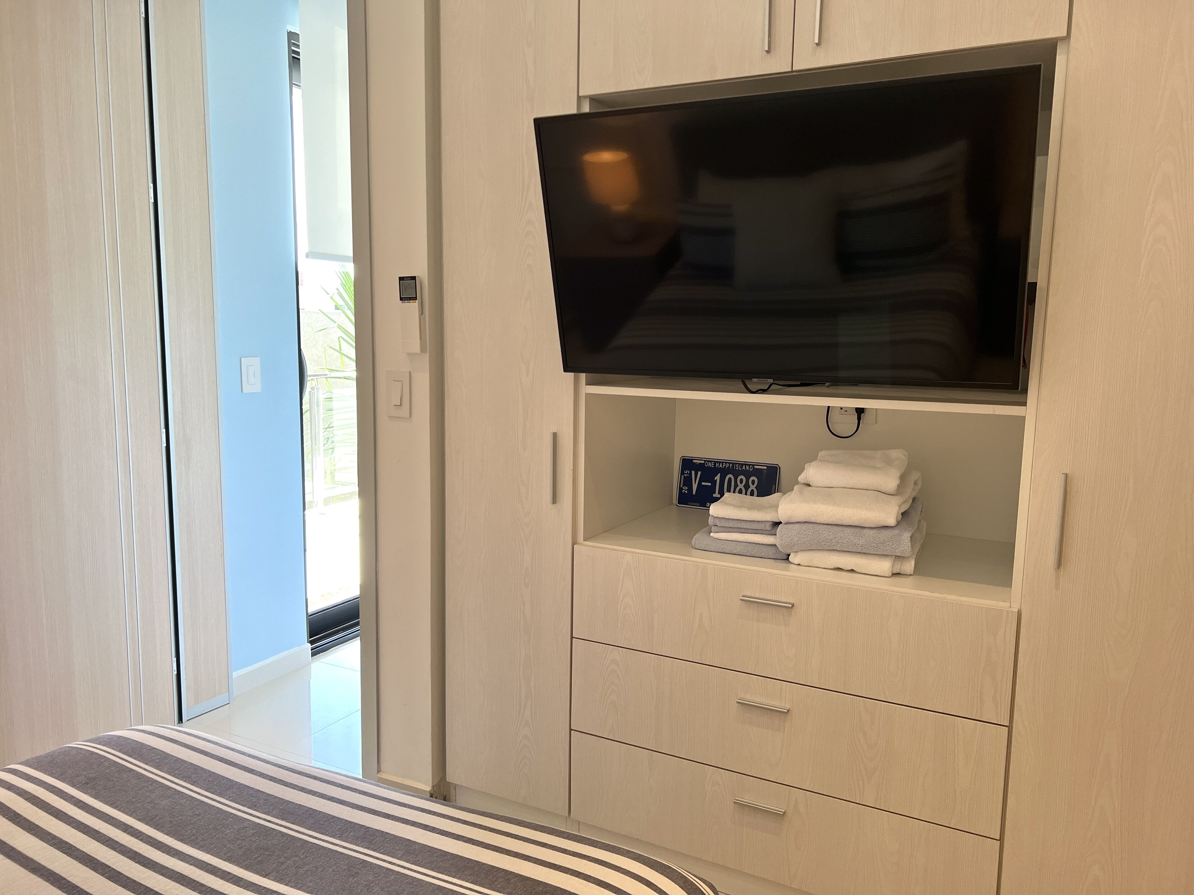 A bedroom with a TV mounted on a wooden cabinet, a bed with striped bedding, and a view of the outside.