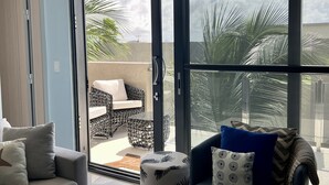 A modern living room with a grey sofa, a blue armchair, and a small round table. A sliding glass door leads to an outdoor patio with wicker furniture and a palm tree. - Waykiri Unit C-12 Luxurious New Apartment (Noord)