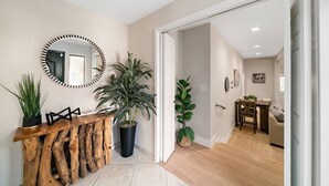 Un interior moderno con una consola de madera, plantas en macetas, un espejo redondo y una zona de comedor con mesa y sillas.