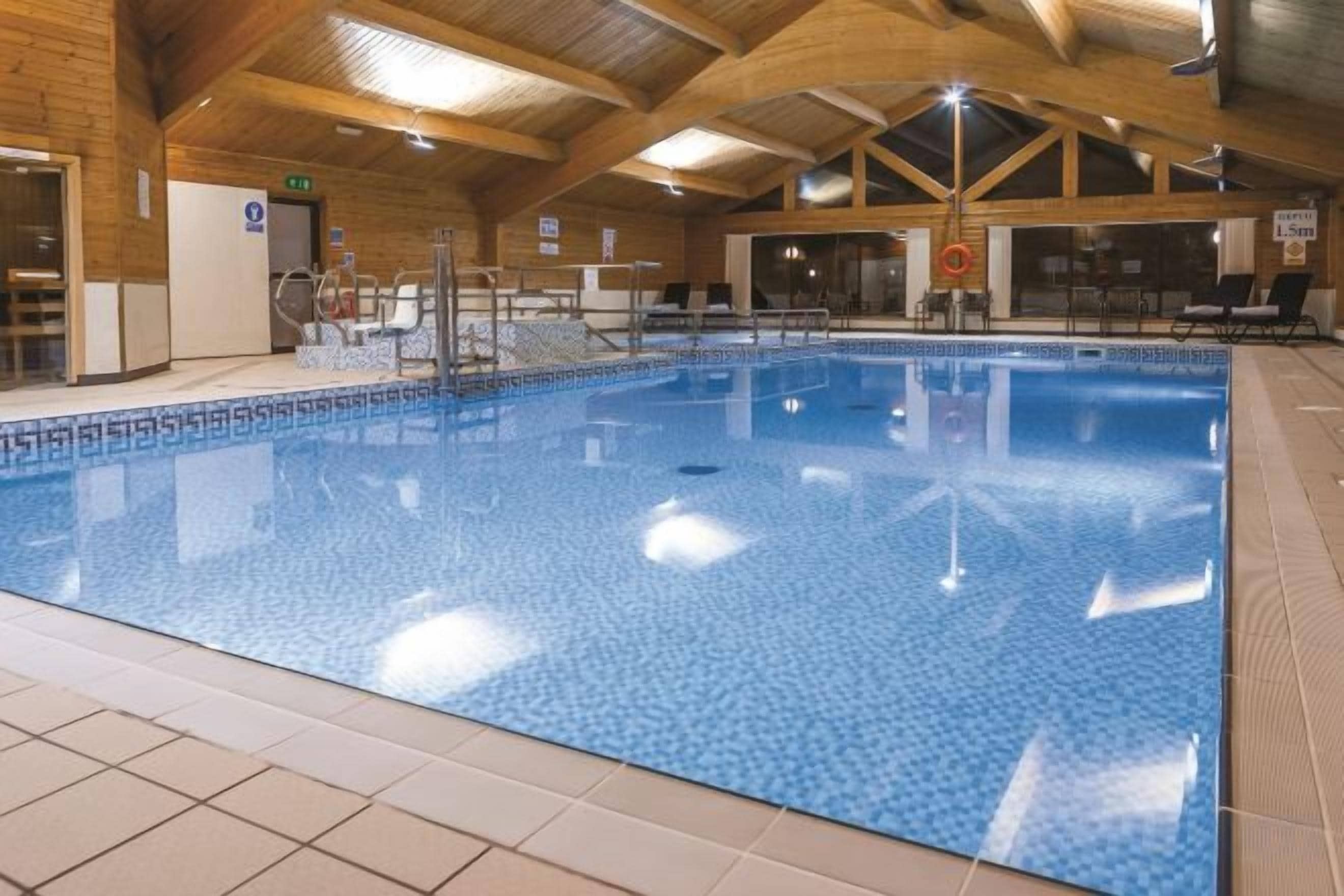 An indoor swimming pool with tiled walls and ceiling, surrounded by lounge chairs and a wooden ceiling structure.