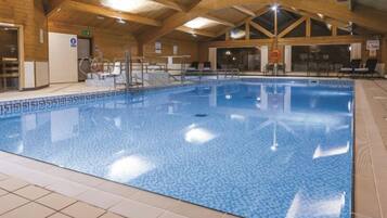 An indoor swimming pool with tiled walls and ceiling, surrounded by lounge chairs and a wooden ceiling structure.