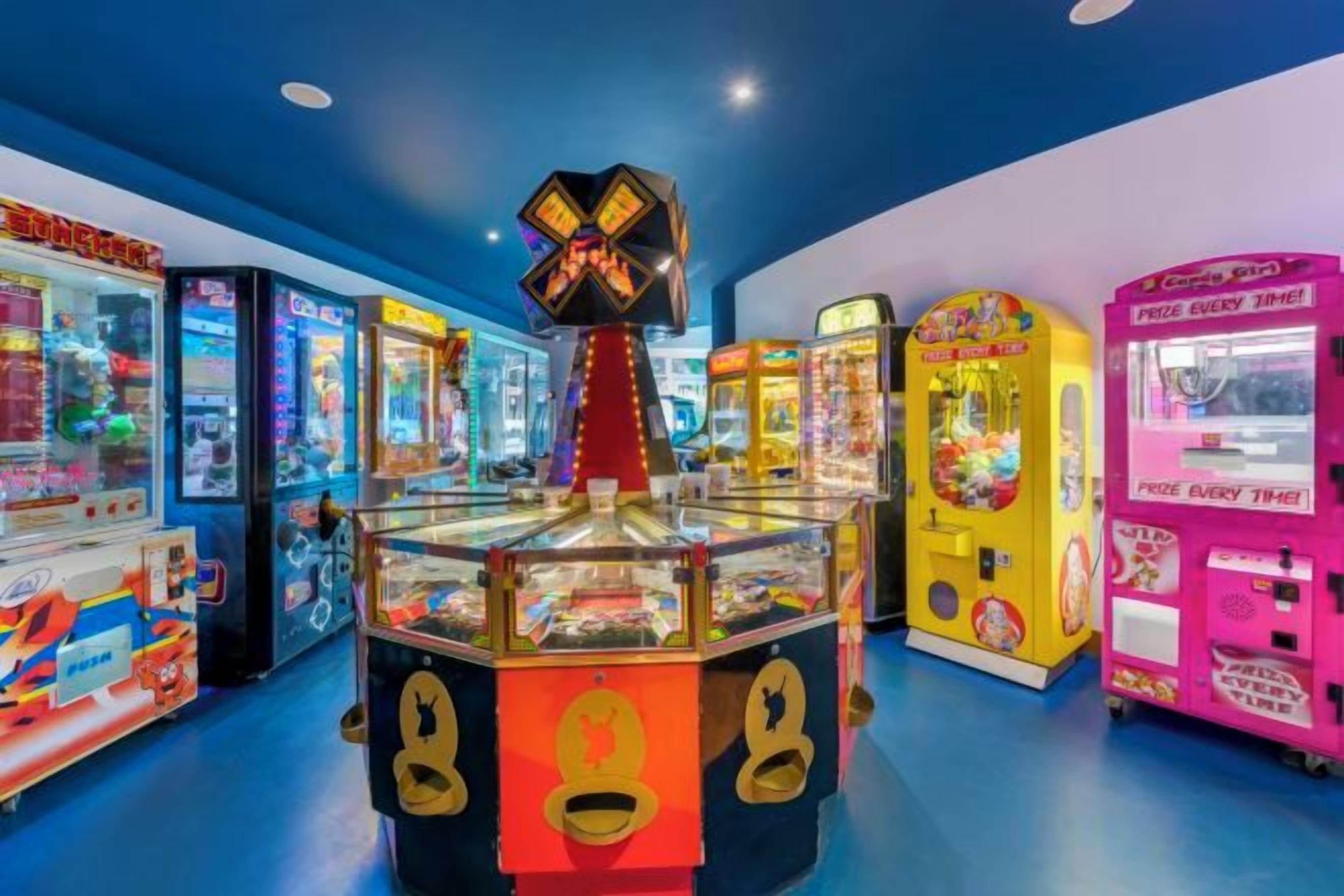A brightly lit arcade with various gaming machines, including claw machines and racing games.