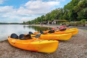 A row of yellow kayaks lined up on a pebbled shore with a calm lake and a building in the background. - Pine Lake Resort (LANCASHIRE)
