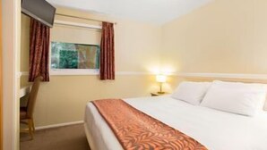 A hotel room with a bed, a desk, a chair, a window with curtains, and a nightstand with a lamp. - Pine Lake Resort (LANCASHIRE)