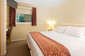 A hotel room with a bed, a desk, a chair, a window with curtains, and a nightstand with a lamp. - Pine Lake Resort (LANCASHIRE)