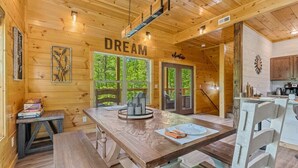 A wooden cabin interior with a dining area, kitchen, and large windows.