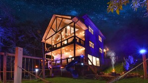 A large, multi-story wooden house with a balcony, illuminated by string lights and a fire pit.