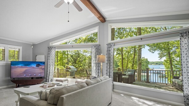 Un salón amplio con un televisor grande, asientos cómodos y un balcón con vistas a un lago y árboles.