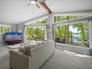 A spacious living room with a large TV, comfortable seating, and a balcony with a view of a lake and trees. - Sheltered Cove - Family-friendly with lots of options! (Jasper)