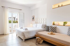 A hotel room with a large bed, a sofa, a balcony with a chair, and a wall-mounted shelf with decorative items. - Mykonaki Hotel (Mykonos)