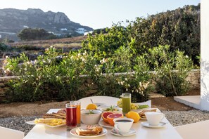 A table set with breakfast items including bread, fruit, and beverages, with a scenic view of mountains and greenery in the background. - Mykonaki Hotel (Mykonos)