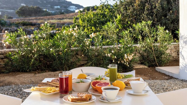 Una tavola imbandita con prodotti per la colazione, tra cui pane, frutta e bevande, con vista panoramica su montagne e vegetazione.