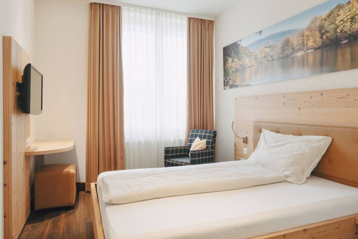 A hotel room with a bed, a chair, a small table, and a painting on the wall.