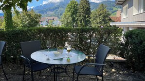 Una mesa redonda con sillas, una copa de vino y un bol de aperitivos, con montañas y un edificio de fondo.