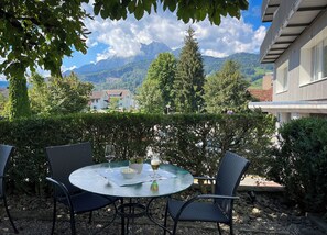 A round table with chairs, a glass of wine, and a bowl of snacks, set against a backdrop of mountains and a building.