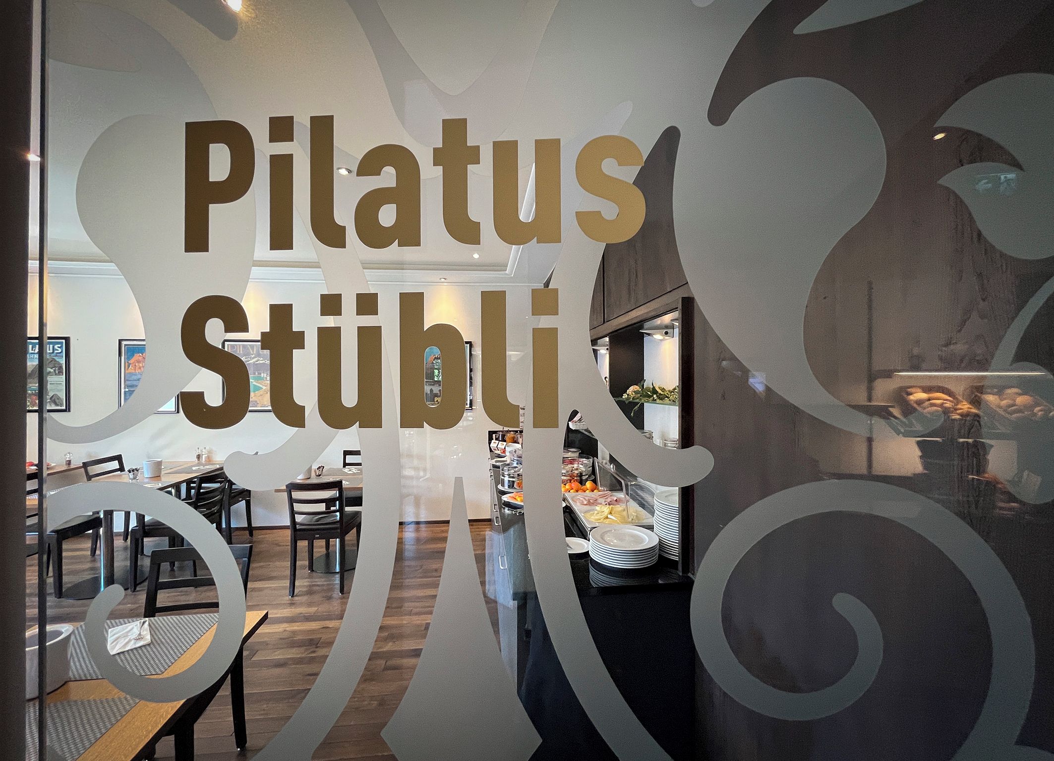 Ein Restaurantinneres mit einer Glastrennwand, auf der 'Pilatus Stübli' steht, und einem Buffetbereich mit Speisen und Getränken.