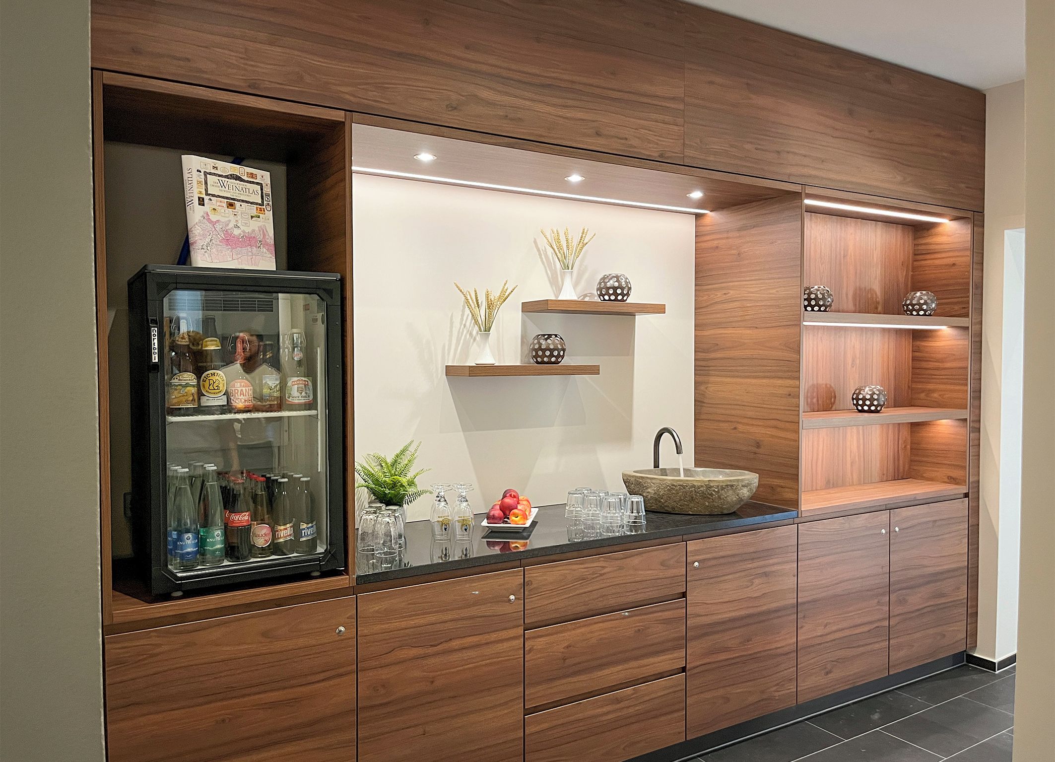 Eine moderne Küche mit Holzausstattung, einem Getränkekühlschrank mit Glastür, einem Waschbecken aus Stein und dekorativen Regalen.