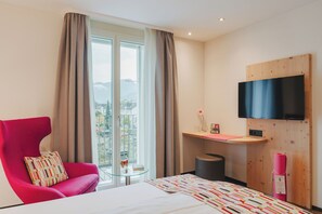 A hotel room with a bed, a red chair, a small table, a TV mounted on the wall, and a view of the outdoors through large windows. - Boutique Hotel Waldegg (Horw)