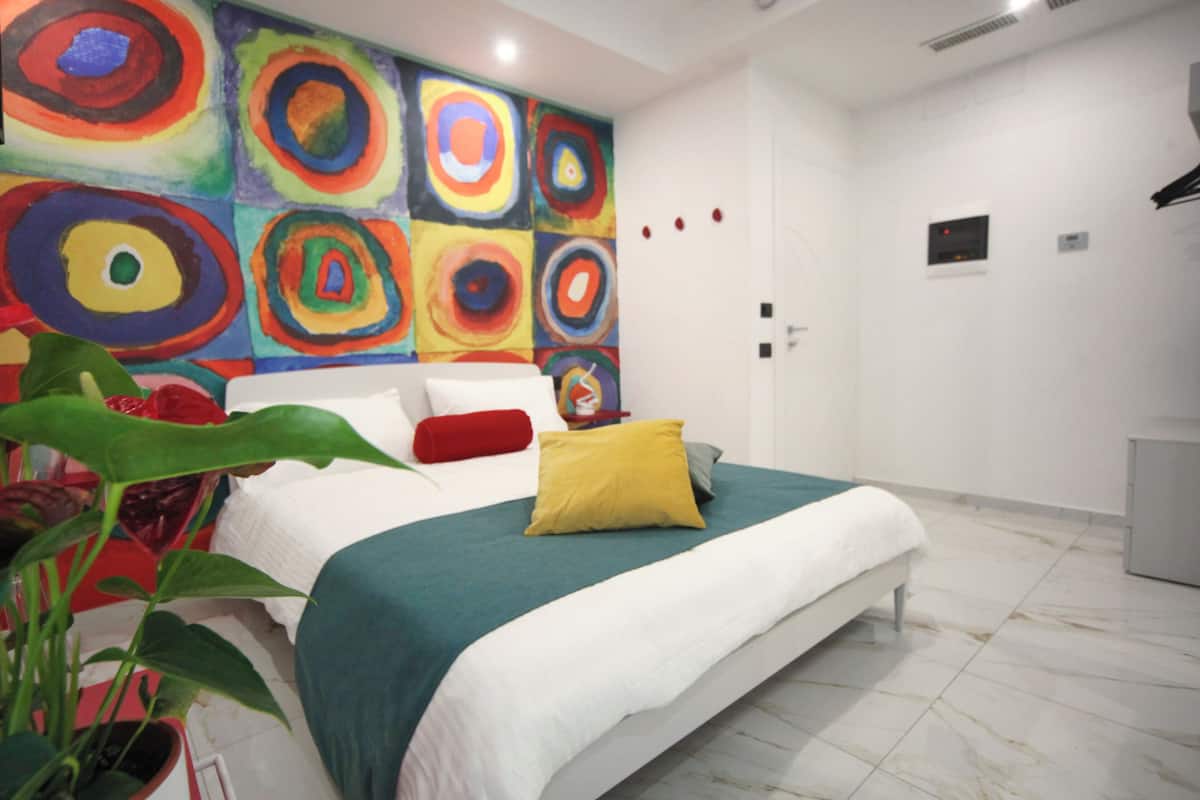 A modern bedroom with a large bed, a colorful abstract painting on the wall, and a potted plant.