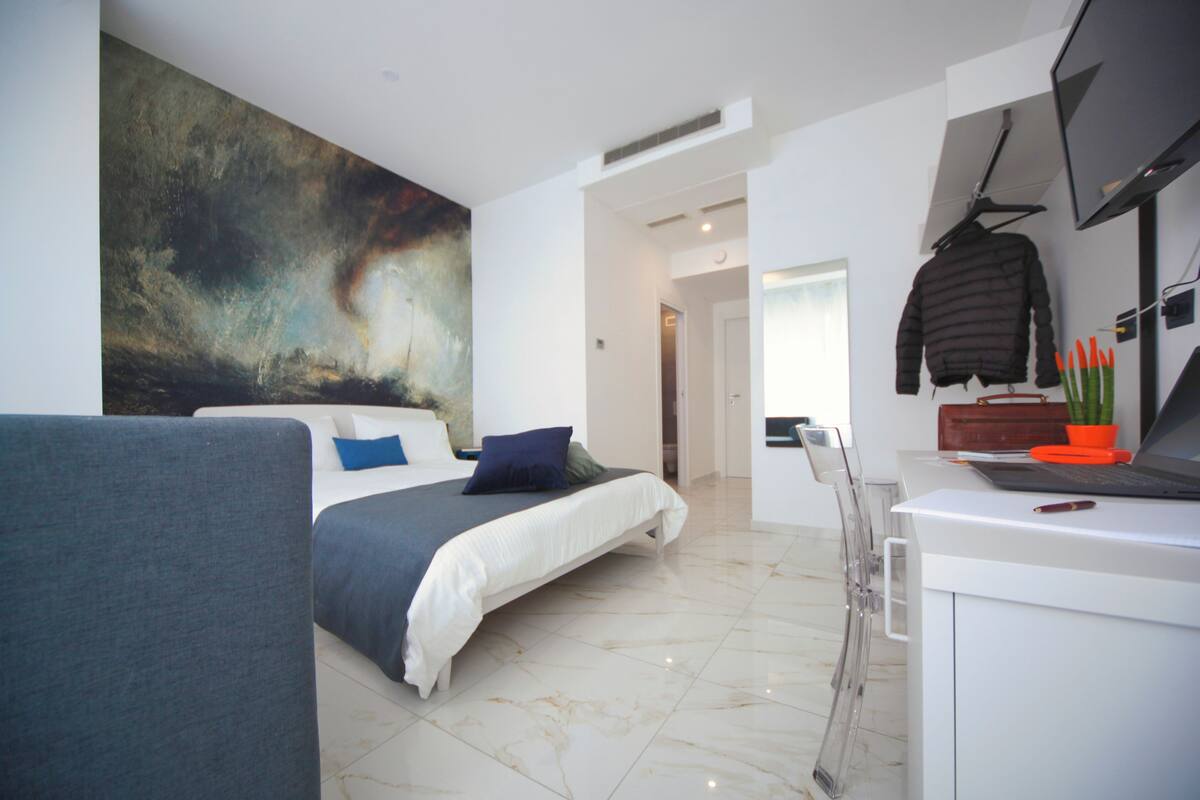 A modern hotel room with a large bed, a desk with a laptop, a TV, and a painting on the wall.