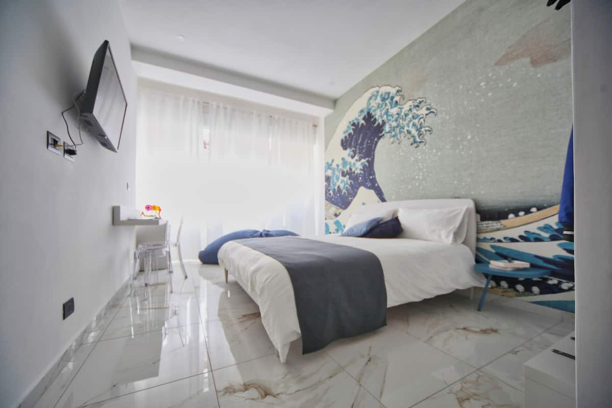 A modern bedroom with a bed, a TV mounted on the wall, and a large artwork depicting a wave.
