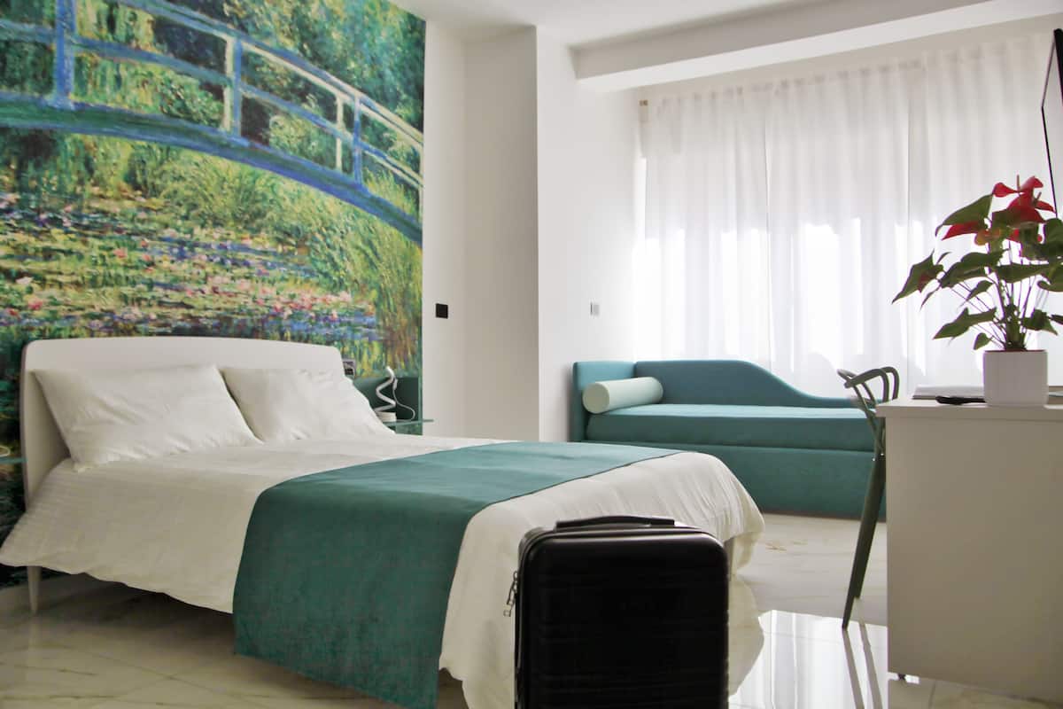 A hotel room with a bed, a desk, a chair, a plant, and a painting of a bridge.