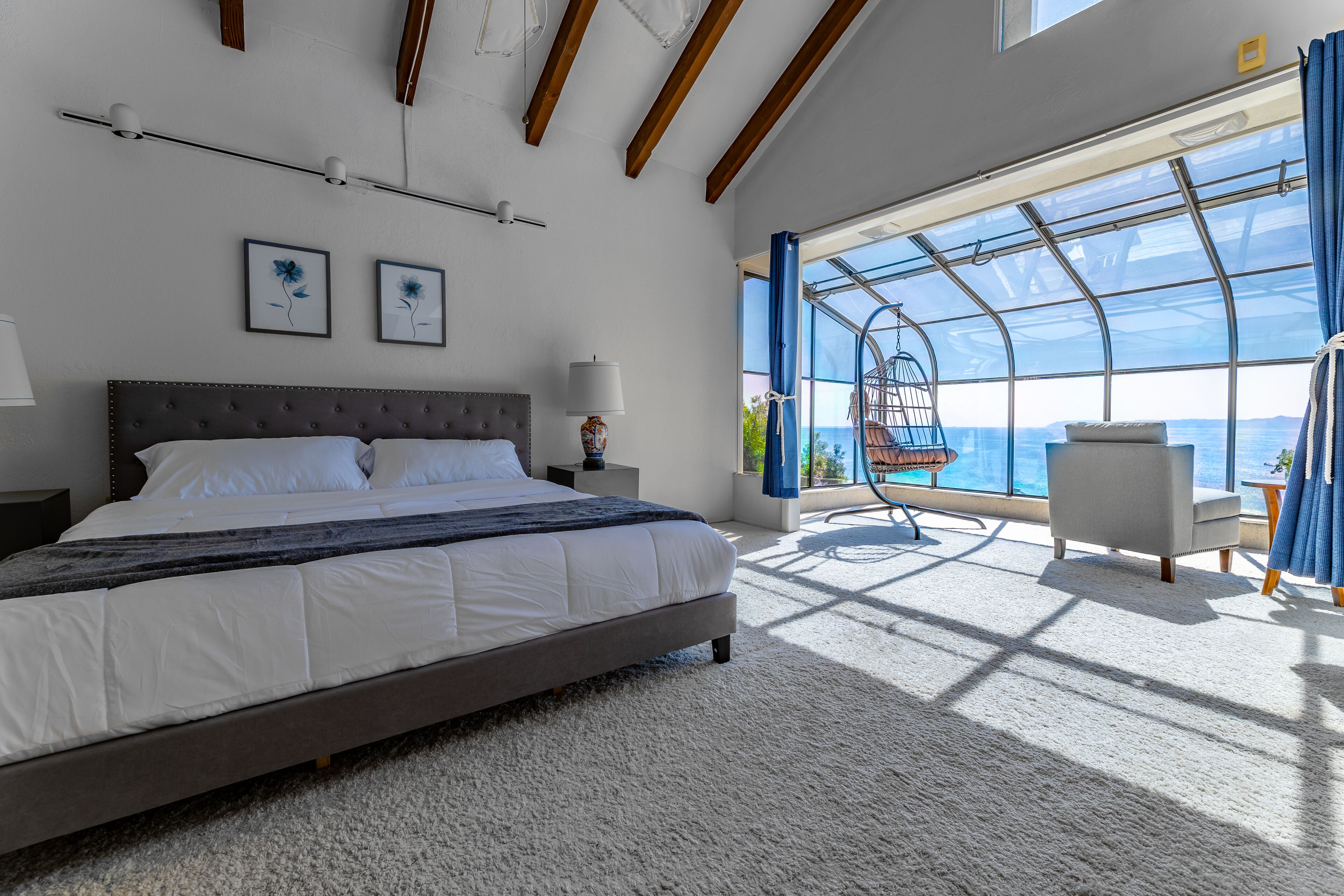 A bedroom with a large bed, a nightstand, a lamp, and a view of the sea through a large window.