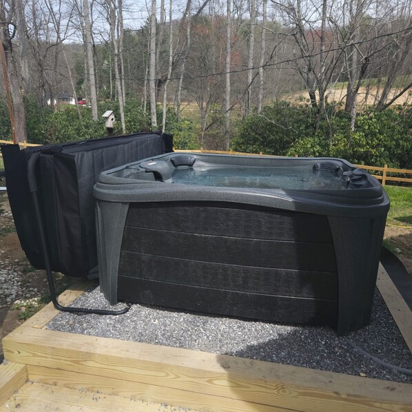 A hot tub in a wooden deck area with a gravel base, surrounded by trees and a wooden fence.