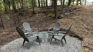 A set of Adirondack chairs arranged around a metal fire pit on a gravel surface, surrounded by trees.