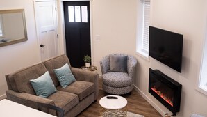 A living room with a brown sofa, a patterned armchair, a small round table, a flat-screen TV, and a fireplace.