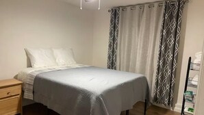Una cama bien tendida con cubrecama gris y almohadas blancas, una mesita de noche de madera con cajón y un carrito metálico con contenedores.