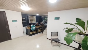 Un hall d'hôtel avec un comptoir de réception portant l'inscription « AYENDA HOTELIER GOLD HOLDING ». Un escalier mène à l'étage supérieur, et on y trouve également un pot de fleurs et une chaise près de la réception.